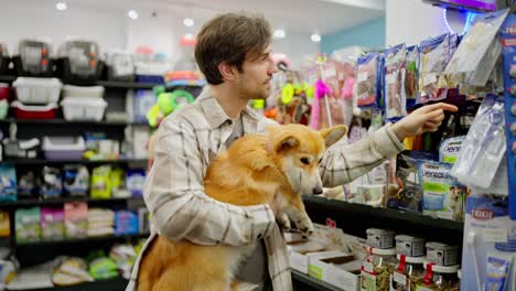 Confident-brunette-guy-holding-his-red-corgi-in-his-arms-while-shopping-and-inspecting-goods-in-a-pet-store