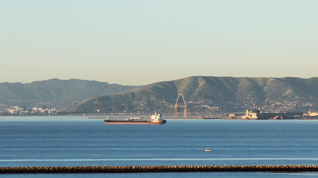 Scenic drone view of a large cargo ship navigating the serene waters of San Francisco Bay, with the industrial port of Oakland and rolling hills creating a beautiful backdrop at sunrise