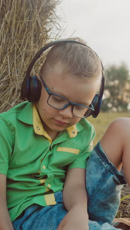 niño con auriculares disfruta de la música mientras está sentado en una manta al aire libre, en un entorno rural tranquilo con una bola de heno en el fondo, un momento pacífico en el campo