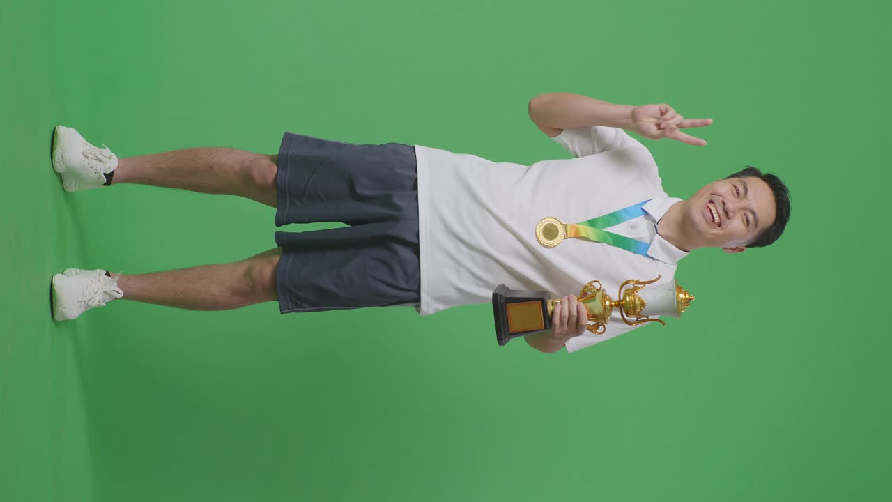 황금 메달과 트로피를 들고 있는 아시아 남성의 몸은 평화의 몸짓을 보여주며 스튜디오의 녹색 스크린 배경에서 첫 번째 우승자로 카메라에 미소 짓고 있습니다.