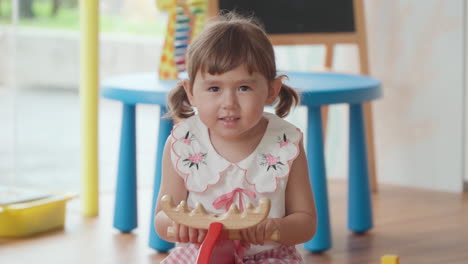 Blonde-Toddler-Girl-Rides-Wooden-Rocking-Deer-in-Slow-Motion-In-a-Brightly-Lit-Playroom-Looking-At-Camera-With-Toothy-Smile-And-Wave-Hand