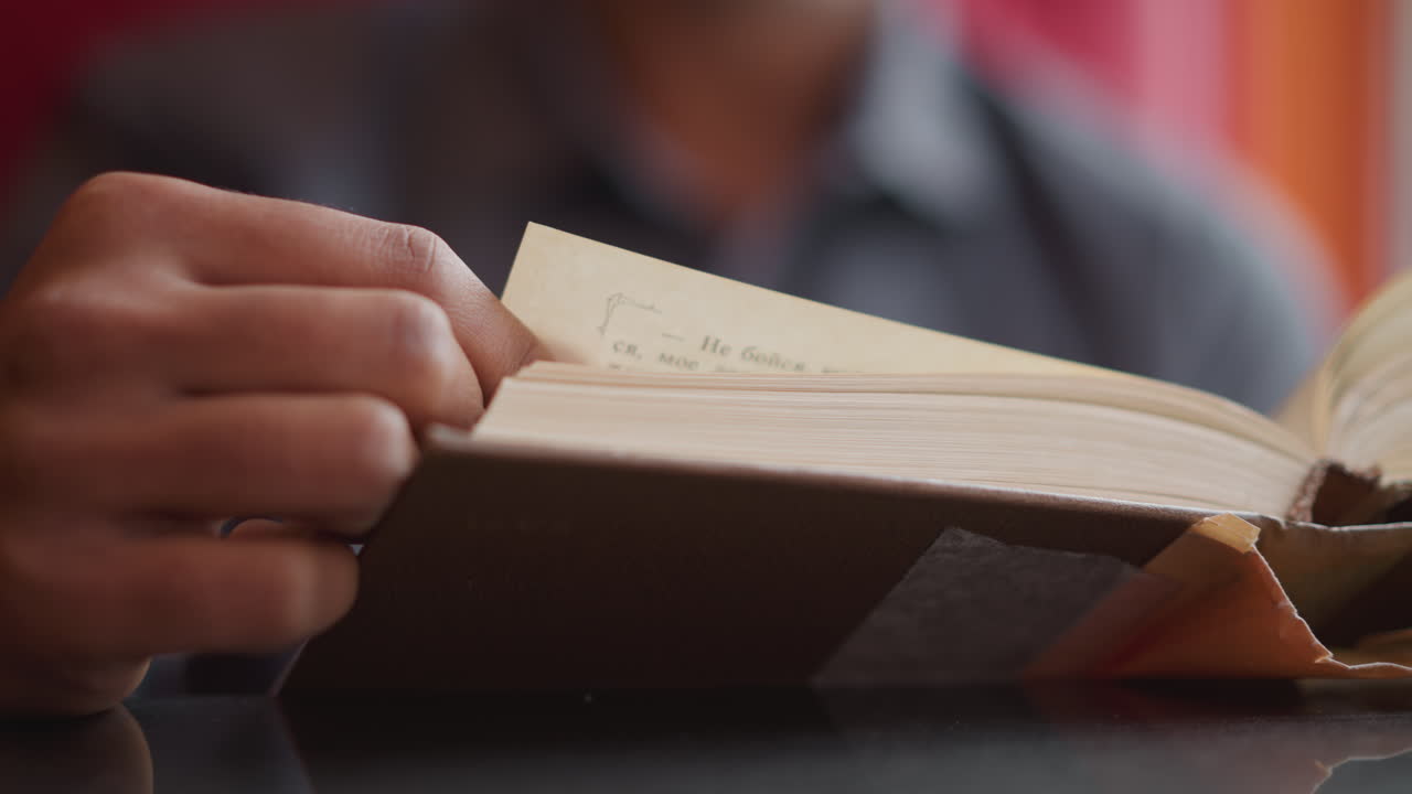 Close-up of hand holding and gently turning page of thick open book on dark surface in soft natural light, suggesting quiet moment of study, concentration, learning, or literary engagement in peaceful setting