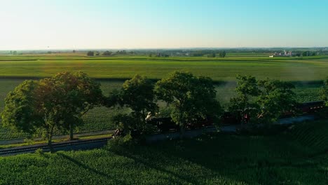 Steam-Train-Passing-through-Amish-Farm-Lands-and-Countryside-on-a-Late-Sunset-Summer-Day-as-seen-by-Drone