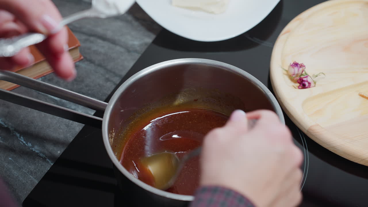 Close-up of cook hand stirring rich, flavorful soup in a pot with a spoon, a cookbook and kitchen equipment are partially visible in the background