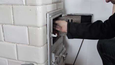 man puts the firewood into old home tiled stove and closes the door