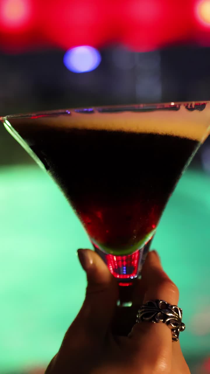 Una mano con anelli mescola un espresso martini in un bicchiere da cocktail, con le luci colorate del bar che si riflettono sulla bevanda, creando movimento dinamico e atmosfera