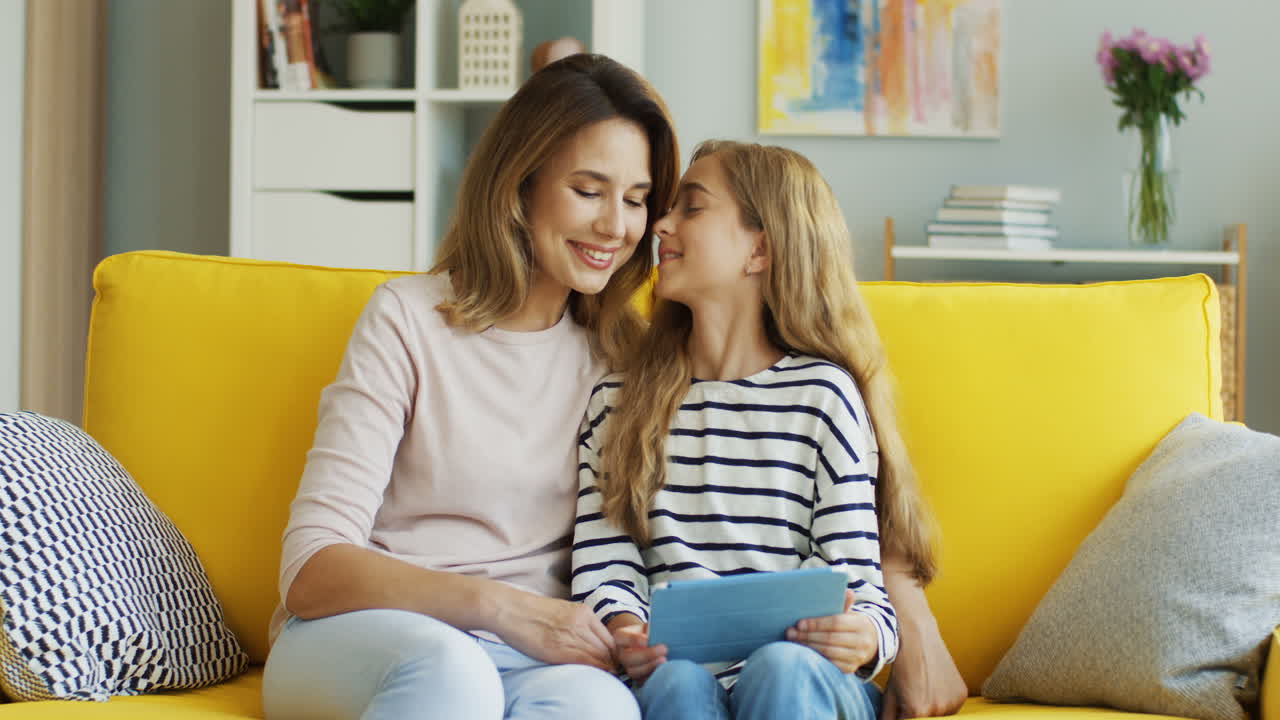 Blonde Mother And Daughter Resting On The Sofa At Home And Watching Something On Tablet