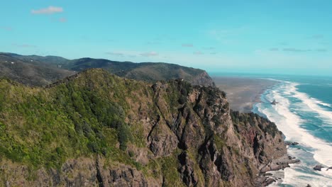 drone-fly-above-wild-nature-rock-cliff-with-blue-clear-sky-and-clean-ocean-water,-relax-calm-tranquil-meditative-scenario-in-Karekare-New-Zealand