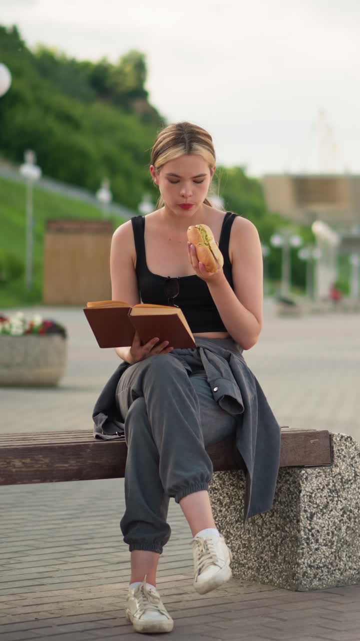 Woman seated outdoors with crossed legs, holding a book in one hand while eating snacks and reading, the blurred background features a lamp post lined up with a blur view of a woman walking