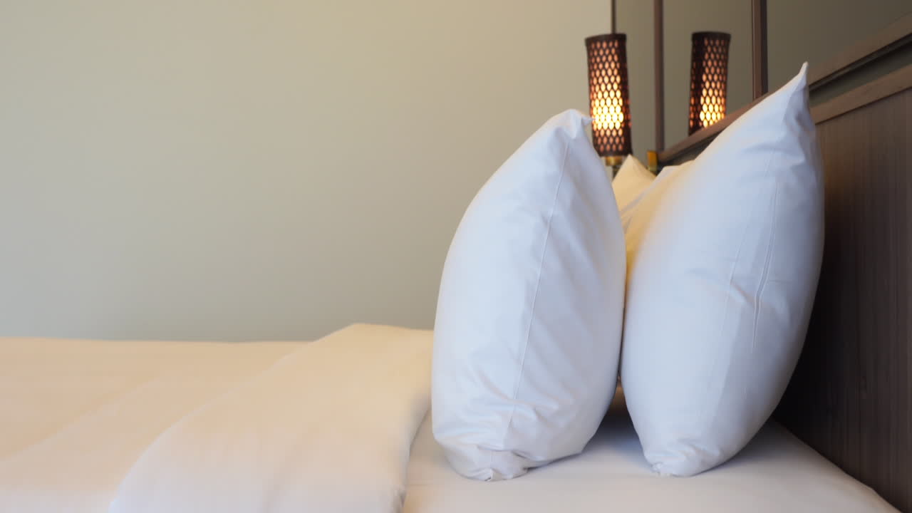 diapositiva verticale della vista laterale di un letto di una camera d'albergo con lenzuola pulite e due strati di soffici cuscini contro la testiera dello specchio e una lampada da parete sullo sfondo