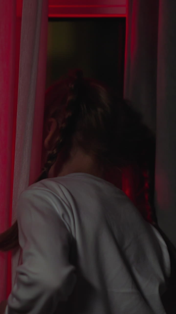 Curious little child with plaits looks out of window attracted by blinking flashlights on street from dark room at night backside view slow motion