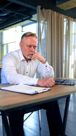 Tired Caucasian male mature businessman sits at desk in the office. Man puts on his glasses and stays focused on his thoughts. Vertical video.