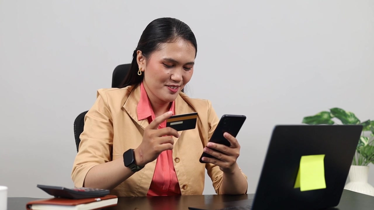 Karyawan wanita bekerja dengan laptop, duduk di meja kerja di ruang kantor sambil memegang kartu kredit dan ponsel.