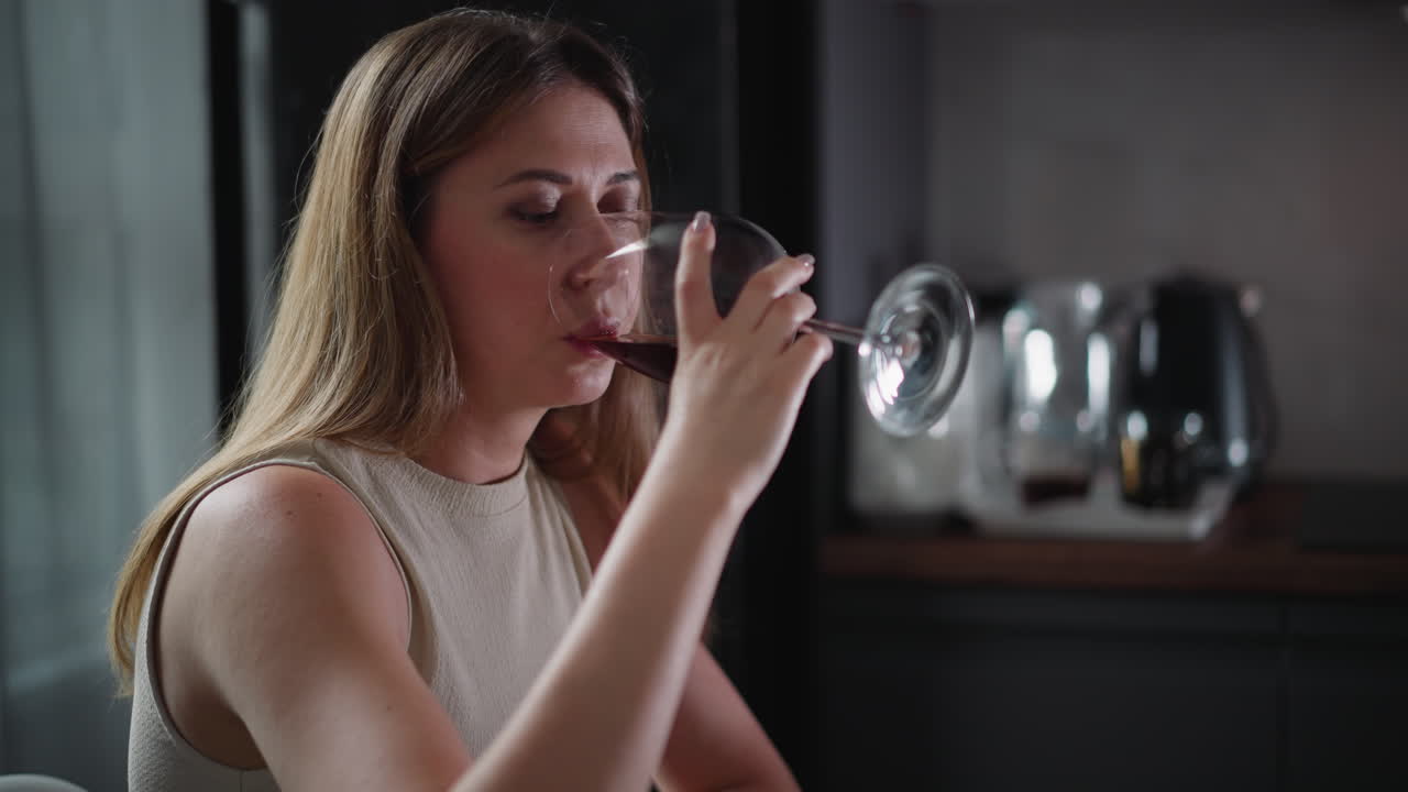 Side view of young woman in sleeveless top sipping red wine from glass with soft blur background, indoor setting with calm lighting, casual yet elegant mood