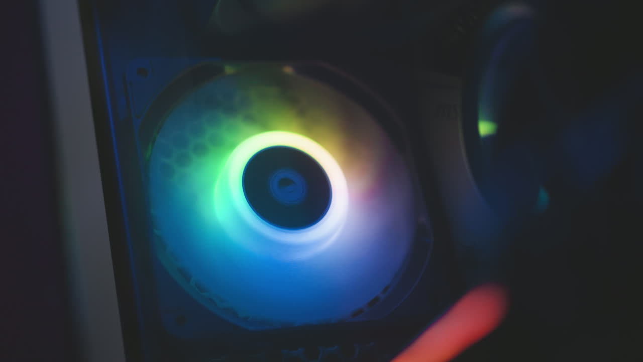Close up of rotating RGB fan glowing with vibrant blue, green, and purple hues inside dark environment, creating smooth ambient lighting effect and colorful reflections on surrounding surfaces