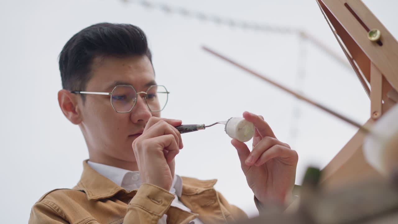 người đàn ông châu Á đang vẽ một bức tượng nhỏ, họa sĩ đường phố phác họa hình thu nhỏ trên giá vẽ, nghệ nhân tập trung phục hồi bình gốm, đeo kính và mặc áo khoác nâu, những nét cọ tinh tế, cận cảnh macro