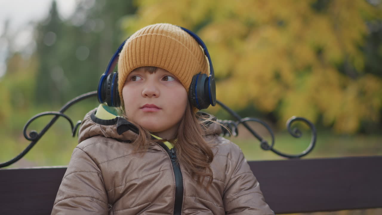 close up little child wearing headphones listening to music outdoors on park bench, thoughtful expression as gentle fingers play with crisp yellow leaf, fall foliage blur backdrop