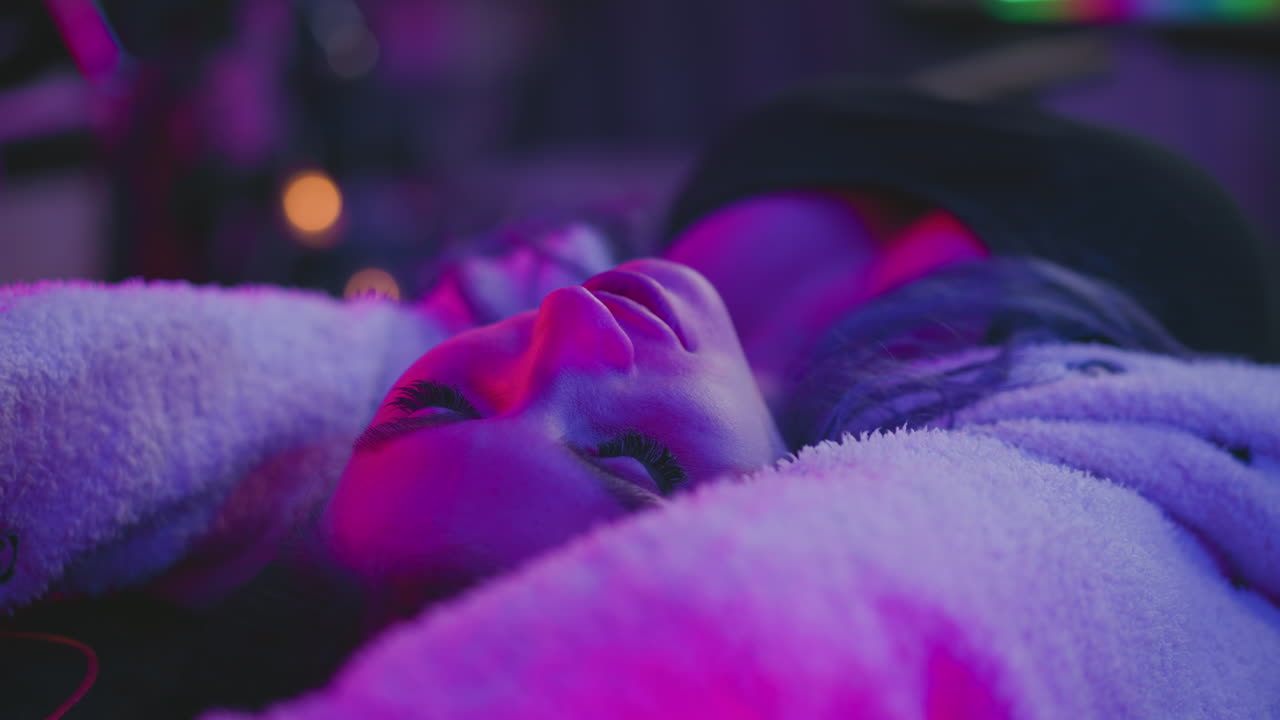 close up of young woman resting with eyes closed while vibrant neon lighting casts dramatic glow over her face and cozy clothing in relaxed tech studio atmosphere during evening session