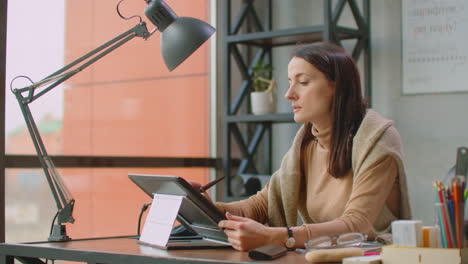 a professional designer in the office draws with a stylus on a graphic tablet sitting in an office with huge windows in the loft style. modern office of graphic designer and creator.