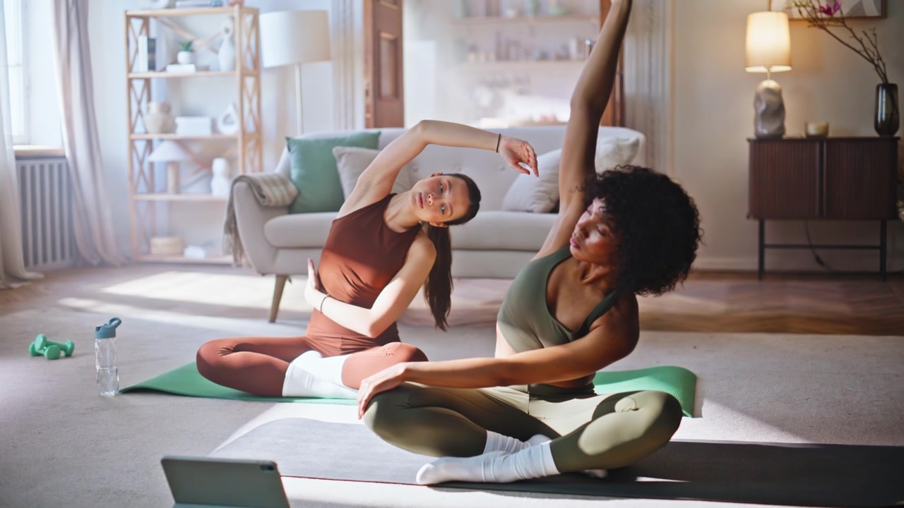 Två kvinnor stretchar yoga i vardagsrummet hemma. Vänner följer ett träningspass på surfplatta.