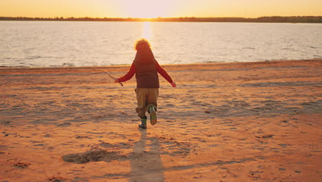 Ein-Glücklicher-Kleiner-Junge-Rennt-Im-Sonnenuntergang-über-Den-Sandstrand-Des-Flusses,-Kindheitsmoment-Und-Glück