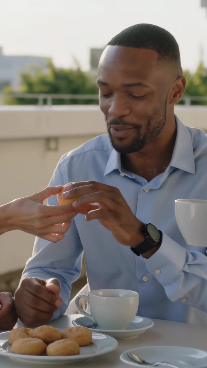 Vertical video: Reaching woman handing pastry to man in blue shirt during rooftop coffee with cups