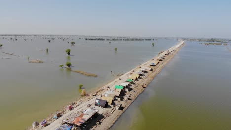 Vista-Aérea-Sobre-Una-Franja-Elevada-De-Tierra-Que-Alberga-Tiendas-De-Campaña-Improvisadas-Rodeadas-Por-Un-Paisaje-Inundado-En-Mehar