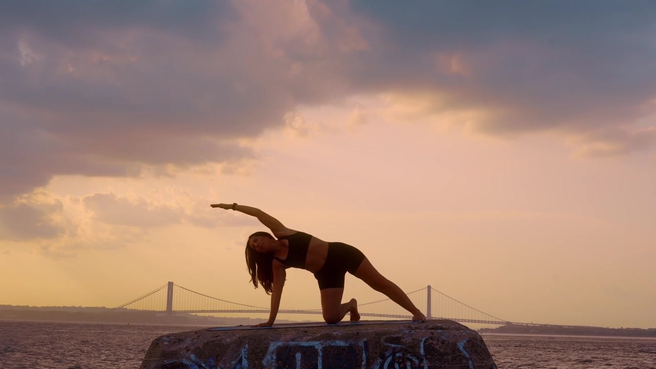 Donna che esegue una posa yoga di allungamento laterale in ginocchio al tramonto a New York. Corpo intero che si piega lateralmente su un molo di pietra con vista sul ponte Verrazzano Narrows.