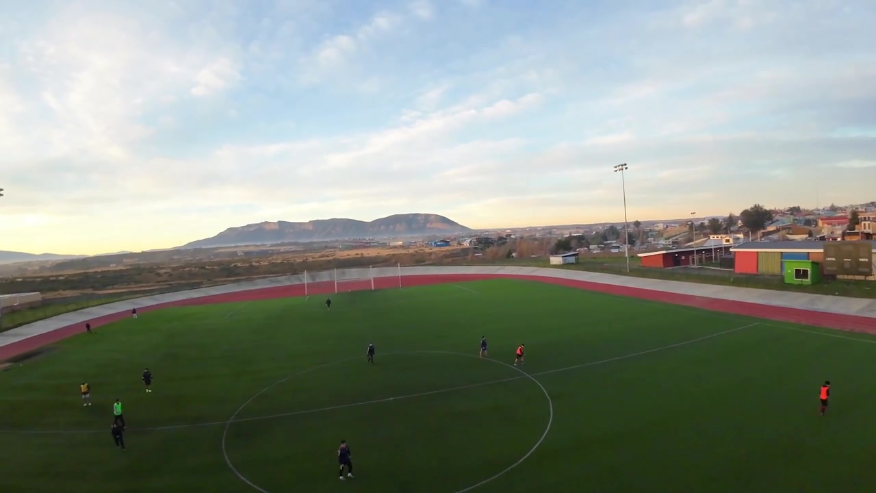 Players training at Estadio Municipal Victor Borquez Miranda surrounded by an athletics track and empty bleachers with mountains and sunset in the distance at Puerto Natales Chile, FPV aerial
