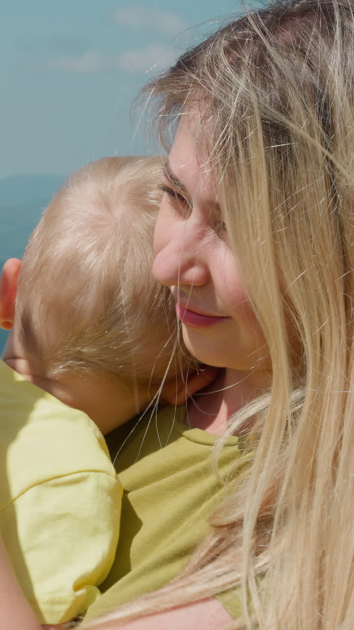 Happy mother with loose dyed fair hair strokes little son leaning on shoulder against old distant mountains at resort closeup slow motion