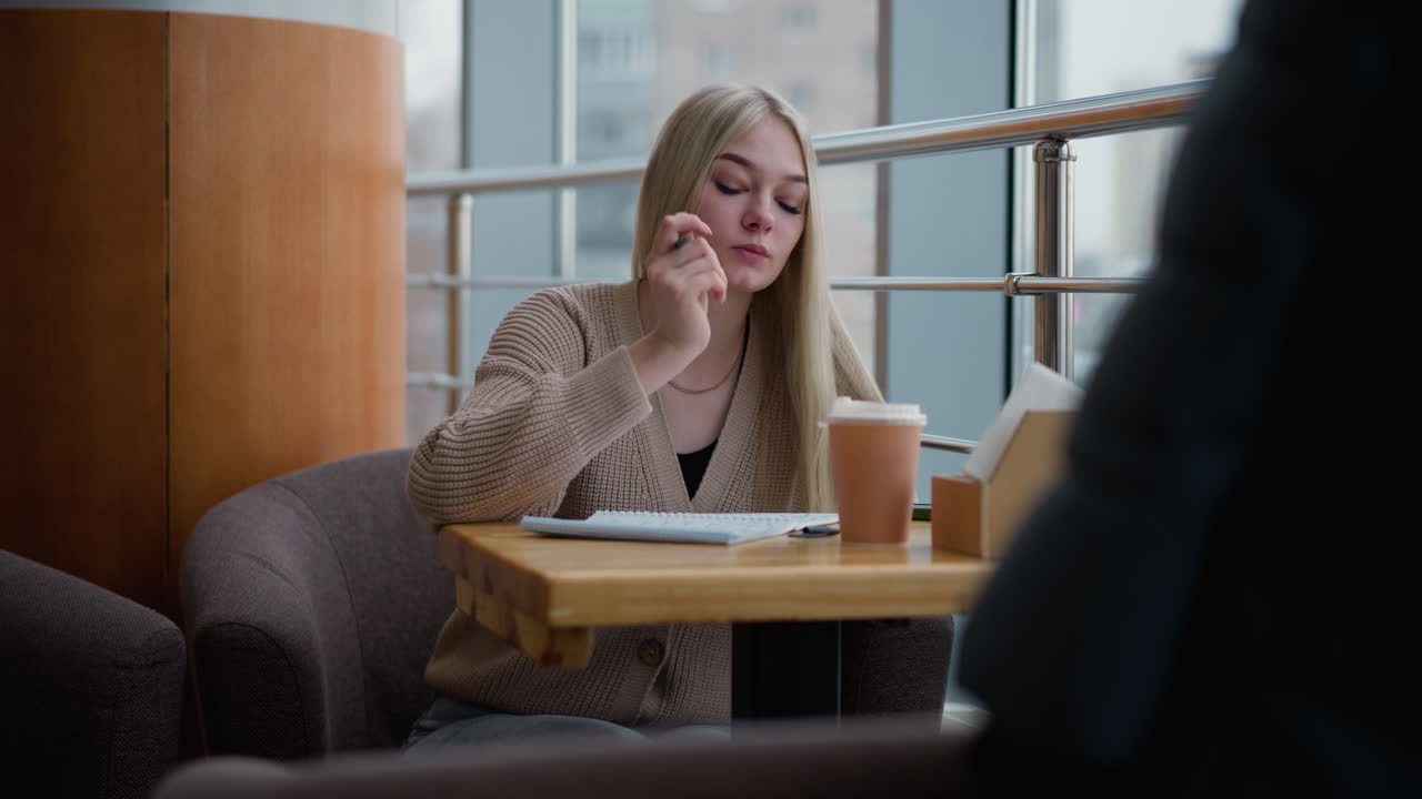 Studerende skriver eftertænksomt på café, kaffekop på bordet, fokuserer på arbejde med glasvinduer, der viser byudsigt i baggrunden, nyder et stille øjeblik med notesbog og kuglepen.