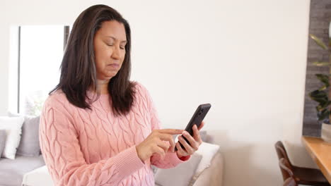 smiling woman in pink sweater using smartphone at home, feeling joyful, copy space