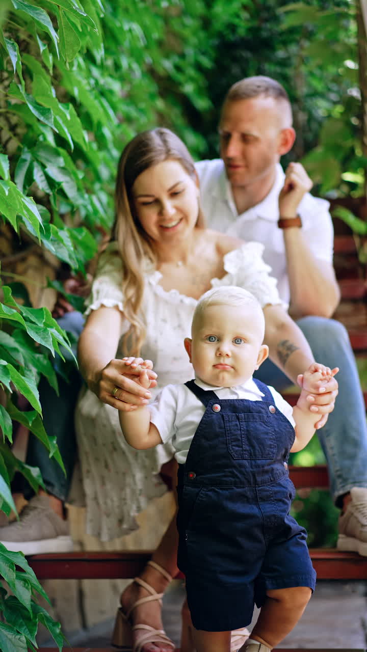 Woman holding by the hands her little baby son wishing to walk ahead. Caucasian family with lovely child outdoors. Vertical video.