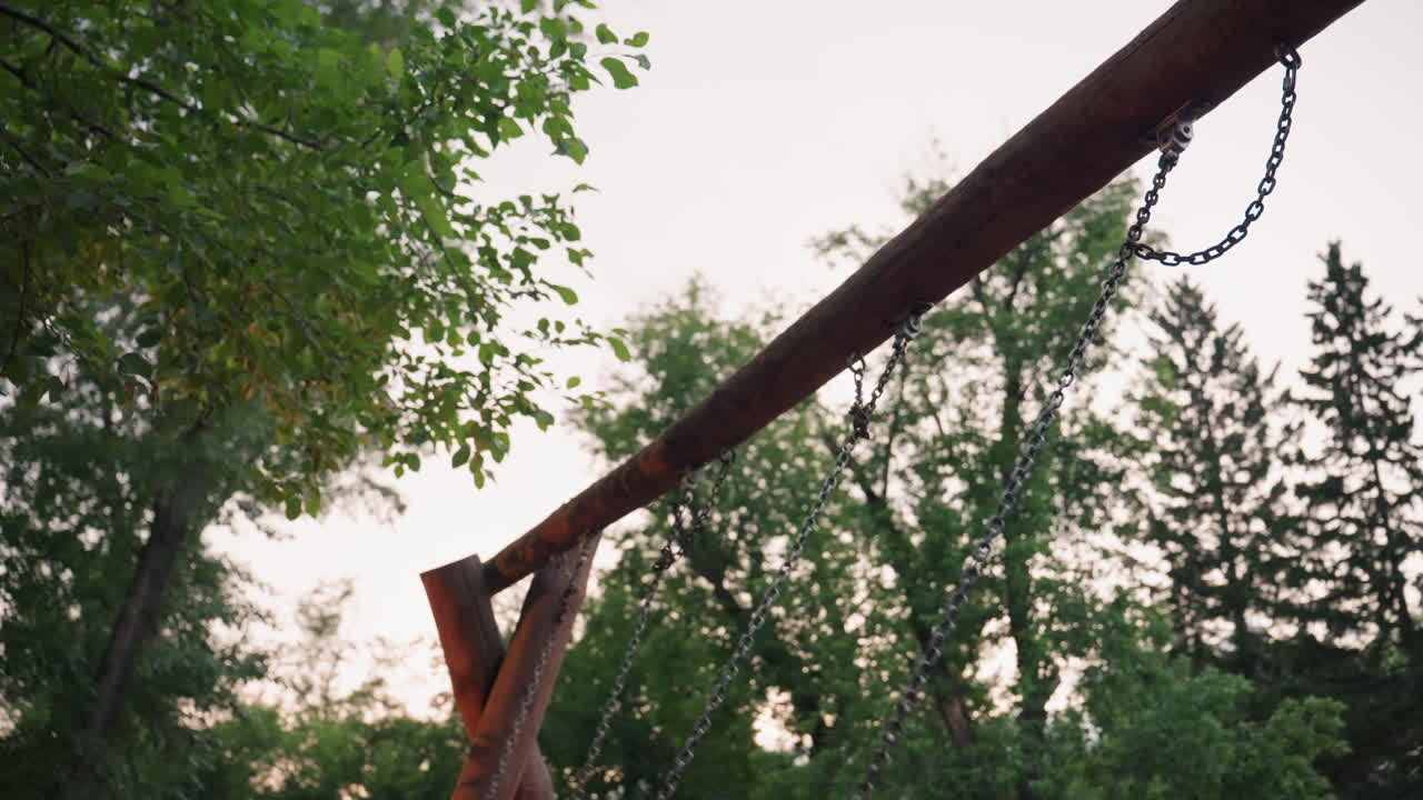 sky view of wooden swing and chains framed by lush green trees at dusk, capturing tranquil playground structure against pale evening sky in serene park setting with soft ambient light