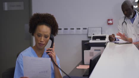 mixed race hospital worker in reception talking on phone with doctors in backgrund