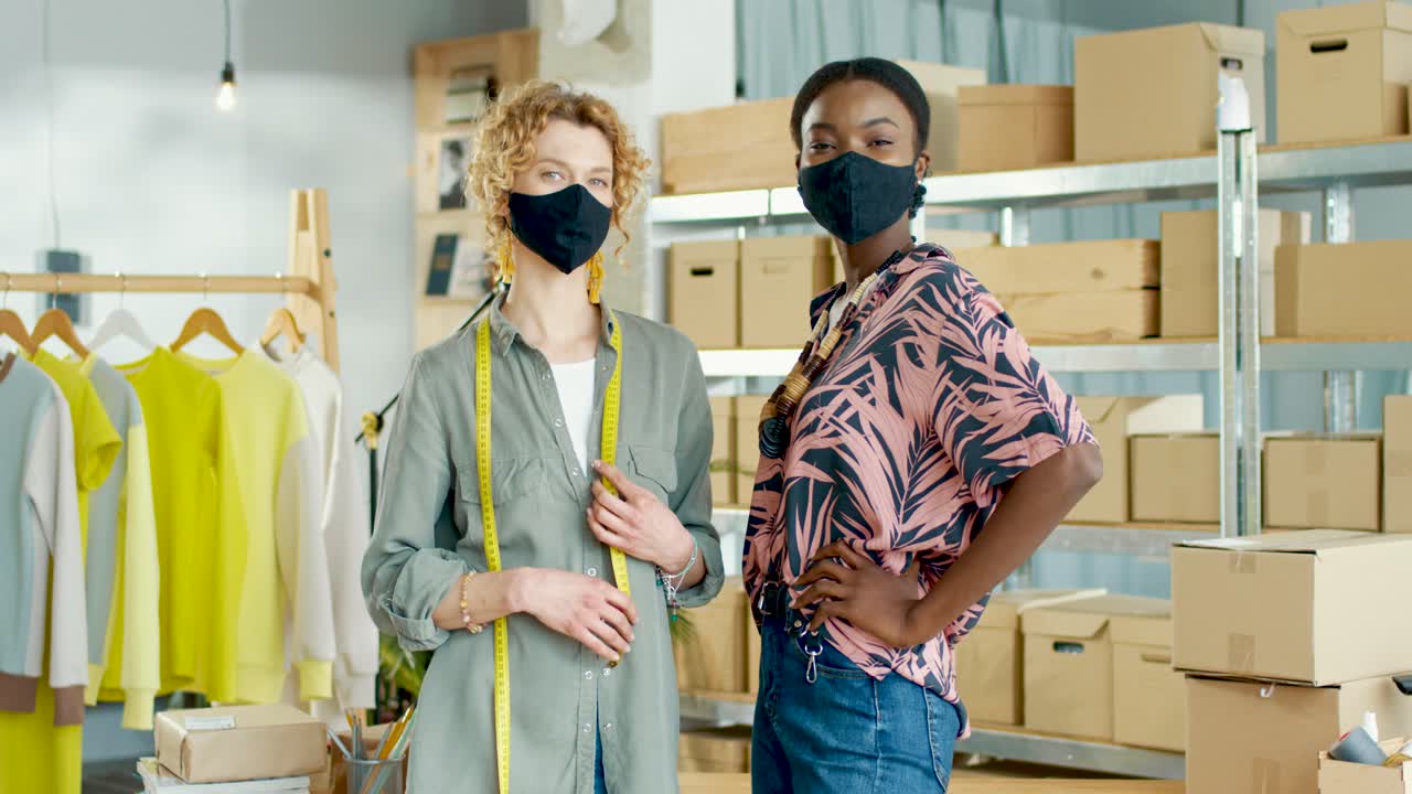 mujeres caucásicas y afroamericanas con máscaras y mirando la cámara en el almacén de la tienda de ropa