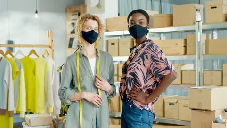 caucasian and african american women wearing masks and looking at camera in clothing shop warehouse