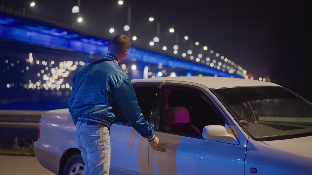 Un uomo bianco sale in auto per un momento di introspezione sotto la luce di un ponte al neon; l'autista siede al volante con uno sguardo intenso e le mani sul volante; riflessi rosa e blu sul viso, cruscotto riflettente, primo piano cinematografico