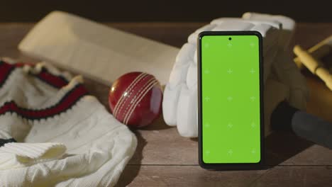 teléfono móvil de pantalla verde rodeado de pelota de bate de cricket y ropa en superficie de madera