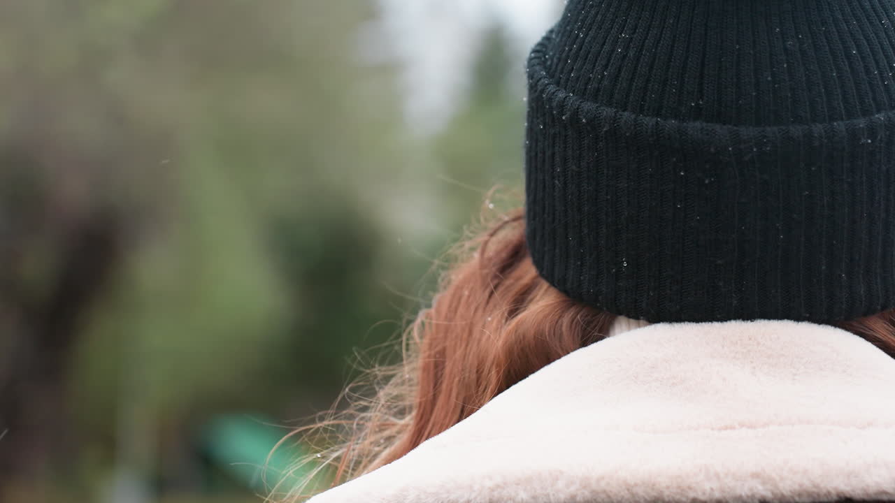 Close back view of student in black knit cap and brown shearling jacket walking outdoors on overcast day, soft focus park and urban background with blurred autumn colors