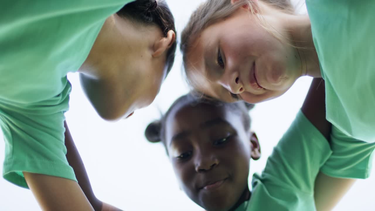 Three teenage female teammates leaning in, linking arms chanting pep talk on field in green shirts