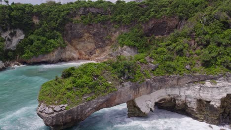 Drone-flies-around-big-green-lush-natural-arch-at-Sumba-island-Indonesia,-aerial