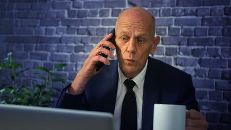 a senior business executive talking on the phone with a coffee in front of a laptop computer in an office