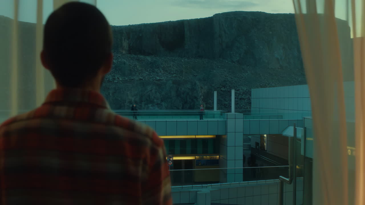Man looks out a window at a modern building complex with people and a rocky mountain in the background