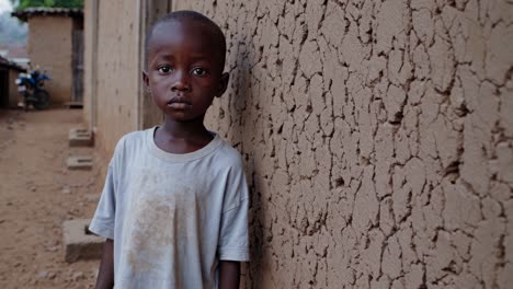 vulnerable african child standing beside weathered mud wall in impoverished rural village, wearing stained clothing and expressing deep emotional isolation through somber expression