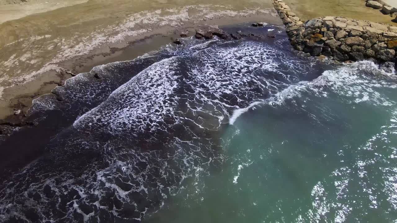 Aerial view of waves crashing onto a narrow beach beside a rocky breakwater. Dark nearshore waters contrast with turquoise sea, highlighting coastal erosion and human shoreline intervention
