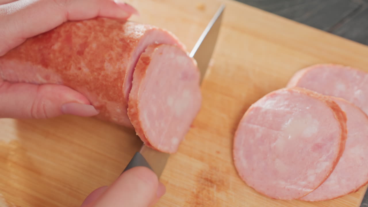 close up hands slicing pink sausage on wooden board using sharp kitchen knife on kitchen counter top under soft lighting showing fresh meat texture and clean preparation in cozy indoor setting