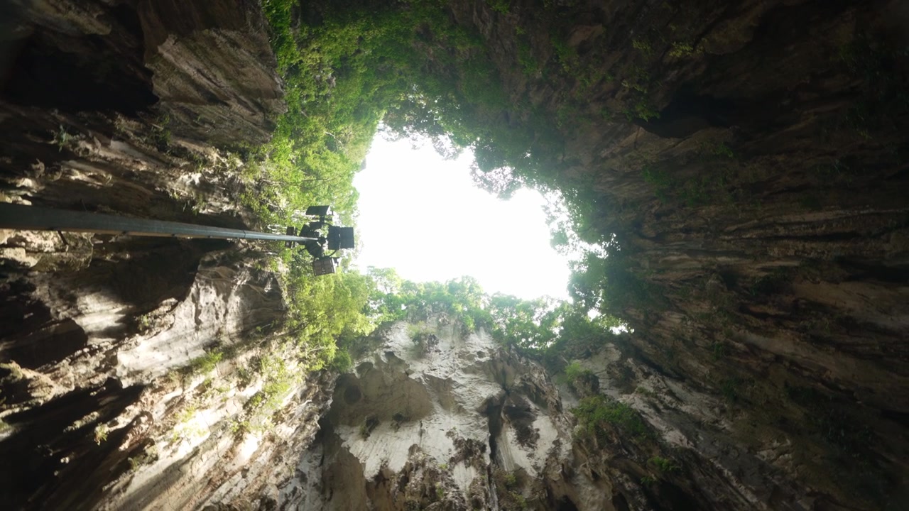 360 rotating shot looking up at Batu Caves opening framed by limestone cliffs and natural light.