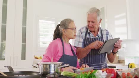 Happy-diverse-senior-couple-using-tablet-and-cooking-in-kitchen