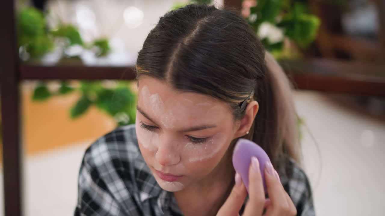 vista de cerca de una mujer joven tocando polvo en su frente usando una esponja de maquillaje púrpura, aplicando polvo translúcido para un acabado liso en su cara mientras prepara el maquillaje